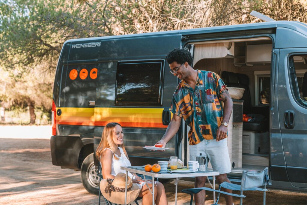 Happy couple having a picnic outside their camper van in a sunny outdoor setting.